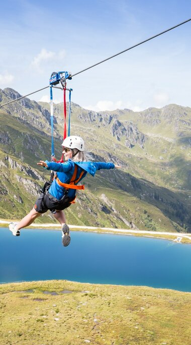 Eine Person gleitet an einer Zipline über einen blauen Bergsee mit beeindruckender Bergkulisse. | © Zillertal Arena