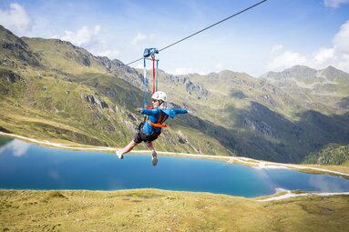 Eine Person gleitet an einer Zipline über einen blauen Bergsee mit beeindruckender Bergkulisse. | © Zillertal Arena