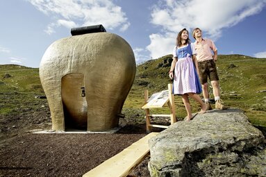 Paar in Trachten vor einer goldenen Glocke am Jodelwanderweg in einer alpinen Landschaft. | © Zillertal Arena