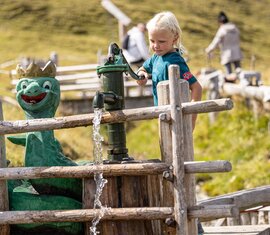 Kind bedient Wasserpumpe neben Drachenfigur auf einem alpinen Spielplatz. | © Zillertal Arena