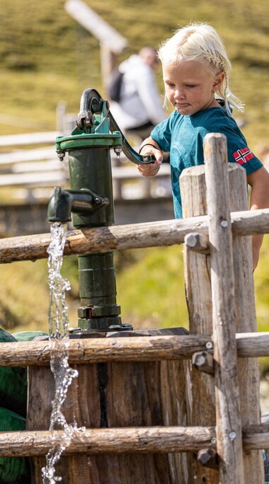 Kind bedient Wasserpumpe neben Drachenfigur auf einem alpinen Spielplatz. | © Zillertal Arena