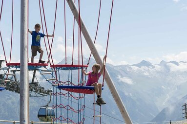 Kinder klettern auf einem Spielplatz in den Bergen mit Seilstrukturen und Gondelbahn im Hintergrund. | © Wildkogel-Arena Neukirchen & Bramberg