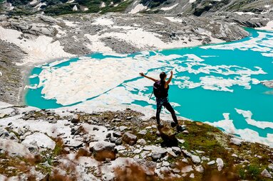 Eine Person steht vor einem türkisblauen Gletschersee, umgeben von dramatischen Berggipfeln. | © Ferienregion Nationalpark Hohe Tauern - Traveloptimizer