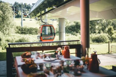 Frühstückstisch mit Aussicht auf eine rote Gondelbahn, eingebettet in grüne Wiesen und Wälder. | © Mittersill Plus GmbH - Daniel Kogler