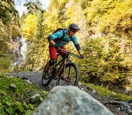 Mountainbiker auf einem Trail mit Wasserfall und herbstlicher Landschaft im Hintergrund. | © Tourismusverband Großarltal