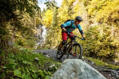 Mountainbiker auf einem Trail mit Wasserfall und herbstlicher Landschaft im Hintergrund. | © Tourismusverband Großarltal