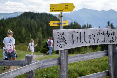 Wegschild von "Tilly's Waldpfad" mit Wanderern im Hintergrund in einer grünen Berglandschaft. | © Tourismusverband Rauris - Lukas Pilz