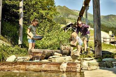 Familie beim Goldwaschen in einem klaren Bergbach mit Holztrögen und sonniger Berglandschaft. | © Tourismusverband Rauris