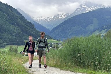 Two hikers on a path through green alpine scenery with mountains in the background. | © Mittersill Plus
