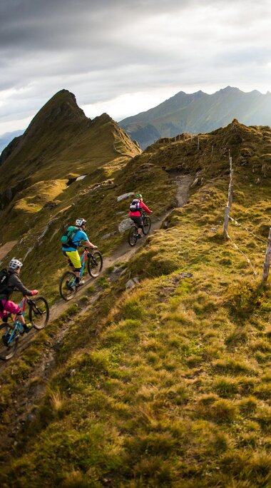 Three mountain bikers ride along a narrow ridge surrounded by alpine scenery. | © SalzburgerLand Tourism