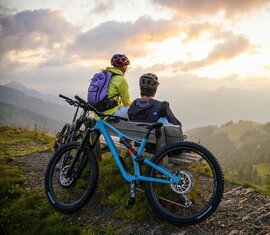 Zwei Mountainbiker genießen den Sonnenuntergang auf einer Bank mit Blick auf das Tal. | © SalzburgerLand Tourismus