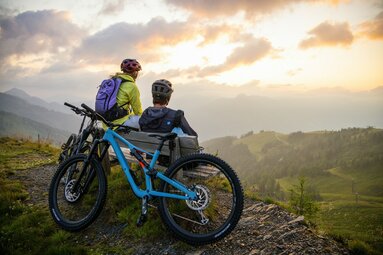 Zwei Mountainbiker genießen den Sonnenuntergang auf einer Bank mit Blick auf das Tal. | © SalzburgerLand Tourismus