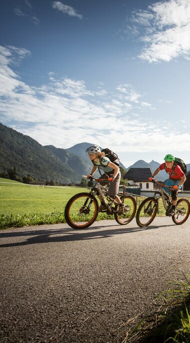Two cyclists on a quiet country road with a church in the background under sunlight. | © SalzburgerLand Tourism