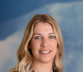 Porträt einer lächelnden Frau mit blonden Haaren in dunkler Kleidung vor blauem Hintergrund. | © Foto Christine
