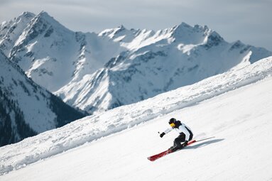 Skifahrer carvt dynamisch eine verschneite Piste mit Bergpanorama. | © Ferienregion Nationalpark Hohe Tauern - Mathäus Gartner