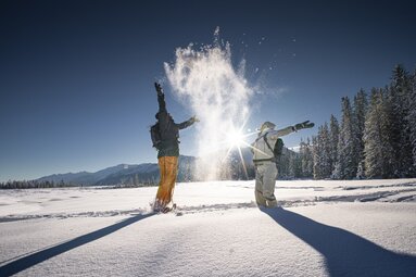 Zwei Menschen werfen Schnee in die Luft im Winterwald bei Sonne. | © Ferienregion Nationalpark Hohe Tauern - Daniel Breuer