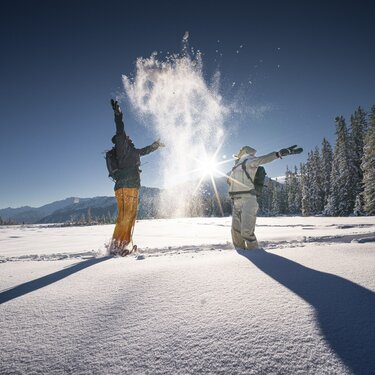 Zwei Menschen werfen Schnee in die Luft im Winterwald bei Sonne. | © Ferienregion Nationalpark Hohe Tauern - Daniel Breuer