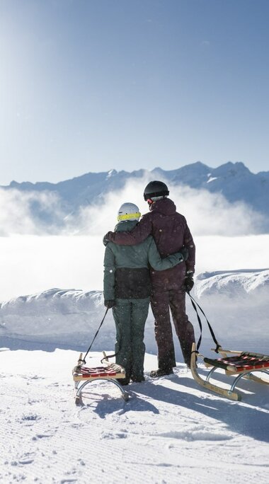 Zwei Personen mit Schlitten blicken auf verschneite Berge und Wolkenmeer. | © Daniel Breuer
