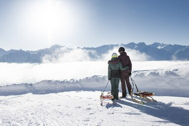 Zwei Personen mit Schlitten blicken auf verschneite Berge und Wolkenmeer. | © Daniel Breuer