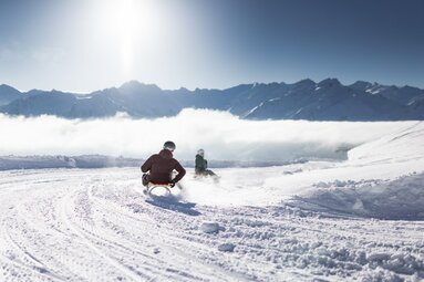 Zwei Personen rodeln auf einer Schneepiste mit Bergpanorama und Sonnenschein. | © Daniel Breuer