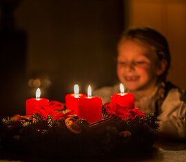 Ein Kind blickt lächelnd auf einen brennenden Adventskranz mit vier roten Kerzen. | © SalzburgerLand Tourismus