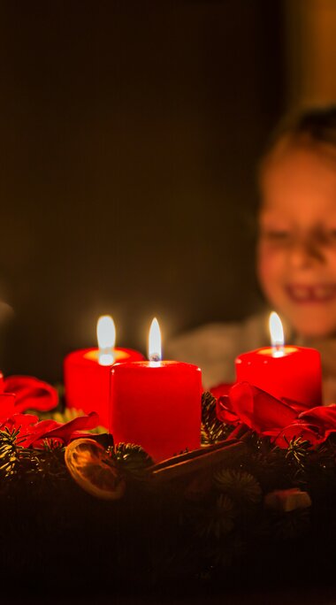 Ein Kind blickt lächelnd auf einen brennenden Adventskranz mit vier roten Kerzen. | © SalzburgerLand Tourismus