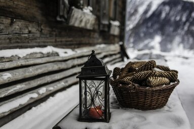 Eine Laterne mit Kerze und ein Korb voller Tannenzapfen stehen im Schnee vor einer Holzhütte. | © TVB Raurisertal - Florian Bachmeier