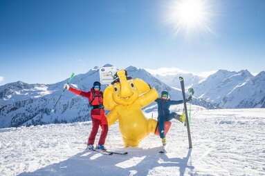 Zwei Skifahrer posieren mit gelbem Elefantenmaskottchen im Schnee vor Bergkulisse. | © Zillertal Arena