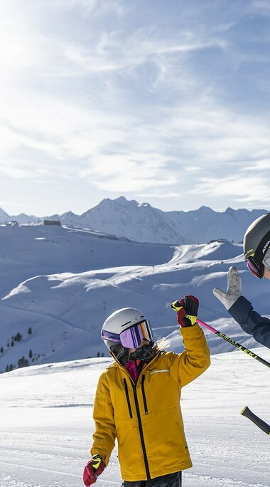 Skifahrer geben sich ein High-Five auf der Piste mit Bergpanorama. | © Wildkogel-Arena Neukirchen & Bramberg