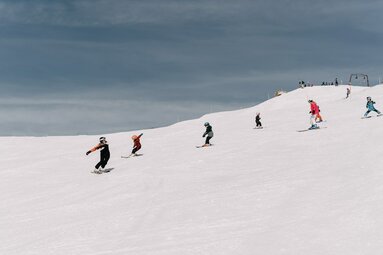  Skifahrer und Snowboarder fahren gemeinsam eine breite Piste bei bewölktem Himmel hinab. | © TVB Uttendorf - Klaus Steiner