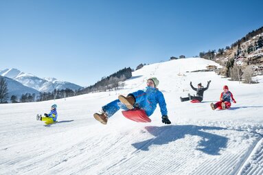 Kinder rodeln fröhlich auf einer verschneiten Piste bei Sonnenschein. | © TVB Piesendorf Niedernsill - Harry Liebmann