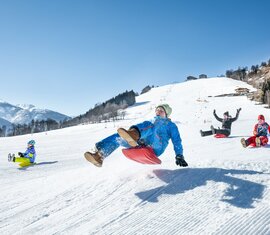 Kinder rodeln fröhlich auf einer verschneiten Piste bei Sonnenschein. | © TVB Piesendorf Niedernsill - Harry Liebmann