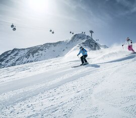 Zwei Skifahrer auf Abfahrt mit Gondeln und Berggipfeln im Hintergrund.  | © Kitzsteinhorn