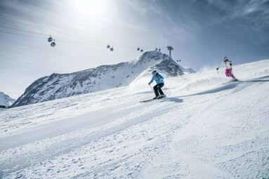 Zwei Skifahrer auf Abfahrt mit Gondeln und Berggipfeln im Hintergrund.  | © Kitzsteinhorn
