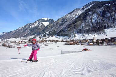 Kind fährt Ski vor verschneitem Dorf und Bergkulisse. | © Tourismusverband BRUCK FUSCH | GROSSGLOCKNER