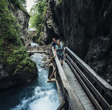 Zwei Wanderer auf einem hölzernen Steg in einer engen, von einem Bach durchflossenen Schlucht. | © Zell am See-Kaprun Tourismus