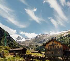 Traditionelle Hütte in einem alpinen Tal mit schneebedeckten Bergen und klarem Himmel.  | © Nationalpark Hohe Tauern - Tobias Kaser