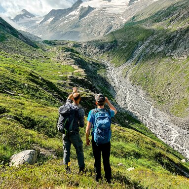 Zwei Wanderer blicken auf ein alpines Tal mit Gletscher und grünen Wiesen. | © Traveloptimizer